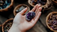 Raw amethyst crystal cluster held in feminine palm against dark background with scattered crystals in wooden bowls, spiritual healing stones and minerals for meditation.