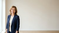 Mature blonde woman in dark blazer and white shirt standing against neutral beige wall near window, professional business portrait for corporate websites and marketing materials.