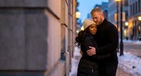 Mature couple embracing on snowy city street at dusk with warm lighting and urban architecture creating romantic winter atmosphere for lifestyle content.