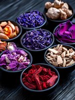 Dried flowers and herbs in black ceramic bowls on dark wooden background. Natural botanical collection of purple, red and pink petals for aromatherapy and decoration.