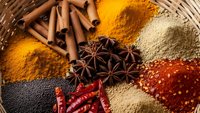 Aromatic spices and seasonings arranged in colorful piles with cinnamon sticks, star anise, and chili peppers on woven basket for culinary cooking.