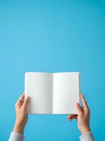 Hands holding open blank notebook with white pages against bright turquoise background for publishing and education projects.