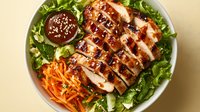 Grilled chicken breast sliced on fresh green salad with shredded carrots and teriyaki sauce in white bowl, served with dipping sauce on beige background.