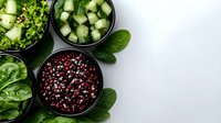 Fresh organic ingredients in black bowls with mixed peppercorns, diced cucumber, and green lettuce leaves on white background, top view food photography.