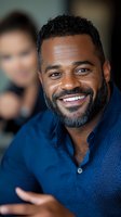 African American man with beard wearing blue button-down shirt smiling warmly at camera. Professional headshot style portrait with soft background blur.