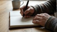 Person writing in an open notebook with a pen at a wooden desk, cozy sweater visible, coffee cup in background, warm tones.