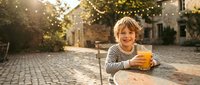 Happy young boy with blonde hair enjoying fresh orange juice at outdoor cafe table on cobblestone street with warm evening lights and cozy atmosphere.