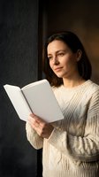 Young Caucasian woman in cream cable knit sweater reading open book by window with warm natural light creating cozy atmosphere for education and leisure.