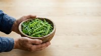 Fresh green peas in wooden bowl held by hands in denim sleeves on light wood surface. Organic produce for cooking and healthy eating.