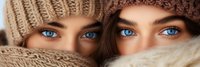 Two young Caucasian women with striking blue eyes wearing cozy knit winter hats and scarves, faces partially covered, winter fashion portrait concept.