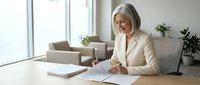 Professional mature blonde businesswoman in beige suit reviewing documents at modern office desk near window with natural light and plants.
