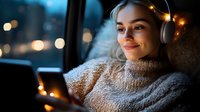 Young woman in cozy sweater wearing headphones and using smartphone at dusk, with warm fairy lights and bokeh creating atmospheric evening mood.