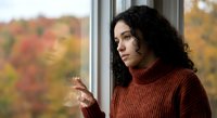 Young woman with curly dark hair in cozy orange sweater looking thoughtfully out window during autumn season with colorful fall foliage background.
