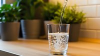 Fresh clean water pouring into transparent glass on wooden table with green plants in background for healthy lifestyle and hydration concept.