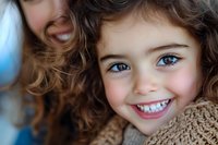Young girl with curly brown hair and bright blue eyes smiling warmly at camera, wearing cozy brown knit sweater. Close up portrait with soft natural lighting.