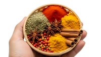 Hand holding wooden bowl filled with colorful spices including turmeric, paprika, cinnamon sticks, star anise and herbs for cooking and seasoning.