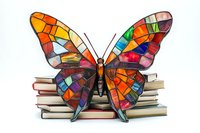 Stained glass butterfly decoration in vibrant colors resting on stack of books against white background, mosaic style wings with geometric pattern.