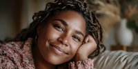 Young African American woman with locs smiling warmly while resting chin on hand in cozy home setting for lifestyle and wellness content.