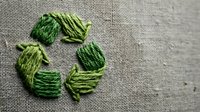 Green embroidery thread arranged in recycling symbol on natural burlap fabric background. Eco-friendly sustainability concept for environmental awareness campaigns.