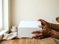 Person with dark skin tone holding white cardboard box on wooden table near window, suitable for e-commerce, delivery, unboxing, and product packaging concepts.
