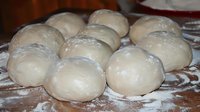 Fresh raw dough balls dusted with flour on wooden surface, ready for baking bread or pizza. Homemade artisanal baking preparation in rustic kitchen setting.