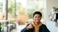 Young man with dark curly hair and medium brown skin tone giving thumbs up gesture in modern office or educational setting, expressing approval and positivity for corporate, business, and success concepts.