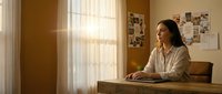 Young Asian woman working on laptop at wooden desk in warm sunlit home office with inspiration board and cozy interior design for remote work productivity.