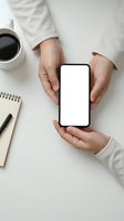 Hands holding smartphone with blank white screen mockup on white desk with coffee cup and notebook for app presentation and mobile interface design.