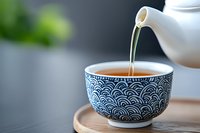 Hot tea pouring from white ceramic teapot into traditional Japanese blue and white wave pattern teacup on wooden coaster against blurred background.
