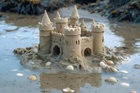Detailed sandcastle with multiple towers and turrets built on beach, decorated with small seashells and pebbles, surrounded by shallow water at shoreline.