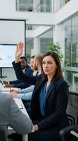 Professional businesswoman raising hand during corporate meeting in modern glass office conference room with diverse team of colleagues.