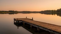 Wooden dock extending into calm lake at sunset with warm orange sky reflecting on water surface creating peaceful lakeside scene for relaxation and nature concepts.