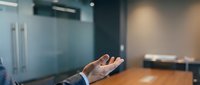 Businessman in formal suit gesturing with open palm during corporate presentation or negotiation in modern office conference room with glass partition and wooden table.