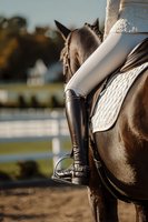 Equestrian rider in black leather boots and saddle on brown horse at outdoor arena, ready for dressage or jumping competition training session.