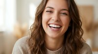 Young Caucasian woman with wavy brown hair laughing joyfully in cozy sweater, expressing genuine happiness and positive emotions in warm indoor setting.