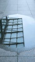 Modern building facade reflected in wet pavement puddle, showing glass windows and metal framework against cloudy sky. Urban architecture photography.