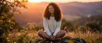 Young African American woman with curly hair sitting cross-legged on rock during golden hour sunset, peaceful meditation pose in nature with mountains background.