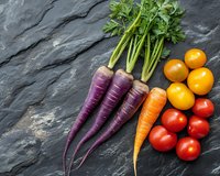 Fresh purple carrots, orange carrot, cherry tomatoes and yellow tomatoes arranged on dark slate stone background. Food photography for healthy eating concept.