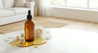 Amber glass dropper bottle with natural skincare oil surrounded by white roses and golden honey on bright wooden floor in modern living room interior.