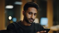 Young South Asian man with beard looking at smartphone in dimly lit cafe, expressing concentration and thoughtfulness while checking messages.