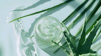 Fresh aloe vera gel in glass jar with natural plant leaves, water drops and soft shadows on light background, organic skincare concept.