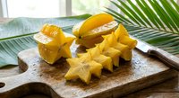 Fresh ripe star fruit slices on wooden cutting board with tropical palm leaves in bright kitchen setting for healthy eating and exotic cuisine concepts.