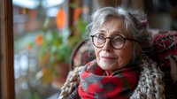 Elegant senior woman with gray hair and glasses wearing red scarf and knit sweater gazes thoughtfully through window, expressing wisdom and contentment in warm natural light.