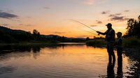 Father and son fishing together at sunset by calm river, enjoying outdoor recreation and bonding time in nature.