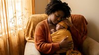 African American mother tenderly holding sleeping newborn baby in cozy home setting with warm natural lighting and comfortable armchair.
