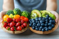 Fresh colorful fruits and vegetables arranged in wooden bowls, including strawberries, blueberries, broccoli, cherry tomatoes, kiwi and avocado, held by hands against gray background.