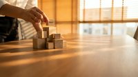 Business professional hand placing wooden blocks in strategic formation on office desk representing growth planning and step-by-step success building.