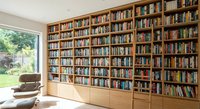 Floor-to-ceiling wooden bookshelf filled with books in modern home library with leather armchair and garden view window for reading and study spaces.