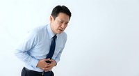 Asian businessman in blue shirt experiencing severe stomach pain holding abdomen with distressed facial expression on white background.