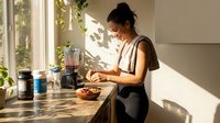 Young Asian woman preparing healthy breakfast in modern kitchen with natural sunlight and green plants creating peaceful morning atmosphere.
